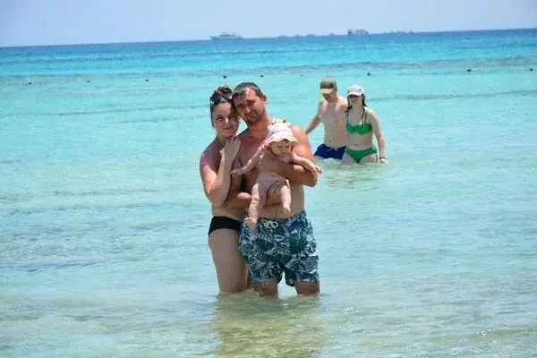 A family of adults and a toddler wading in shallow turquoise water with another couple nearby.