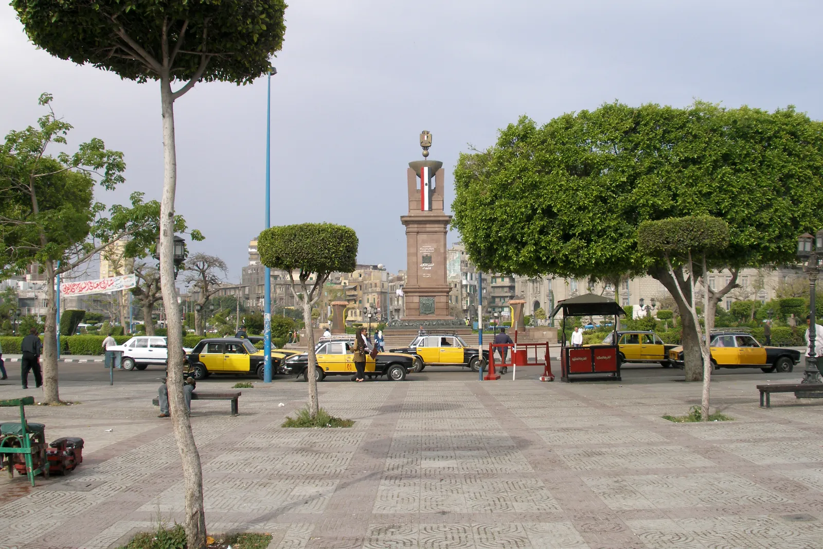 A city square with trimmed trees, a stone monument column, and rows of black-and-yellow taxis parked nearby.