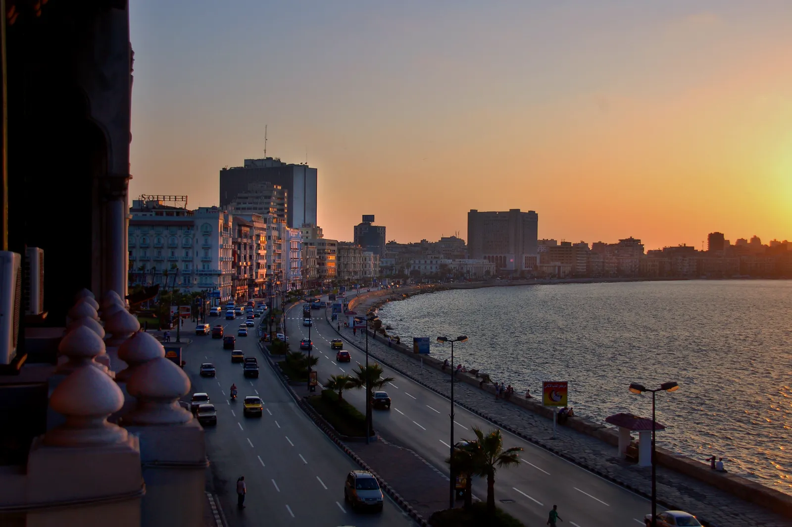 A sunset view over a coastal boulevard with traffic and palm trees, seen from a balcony overlooking the sea.