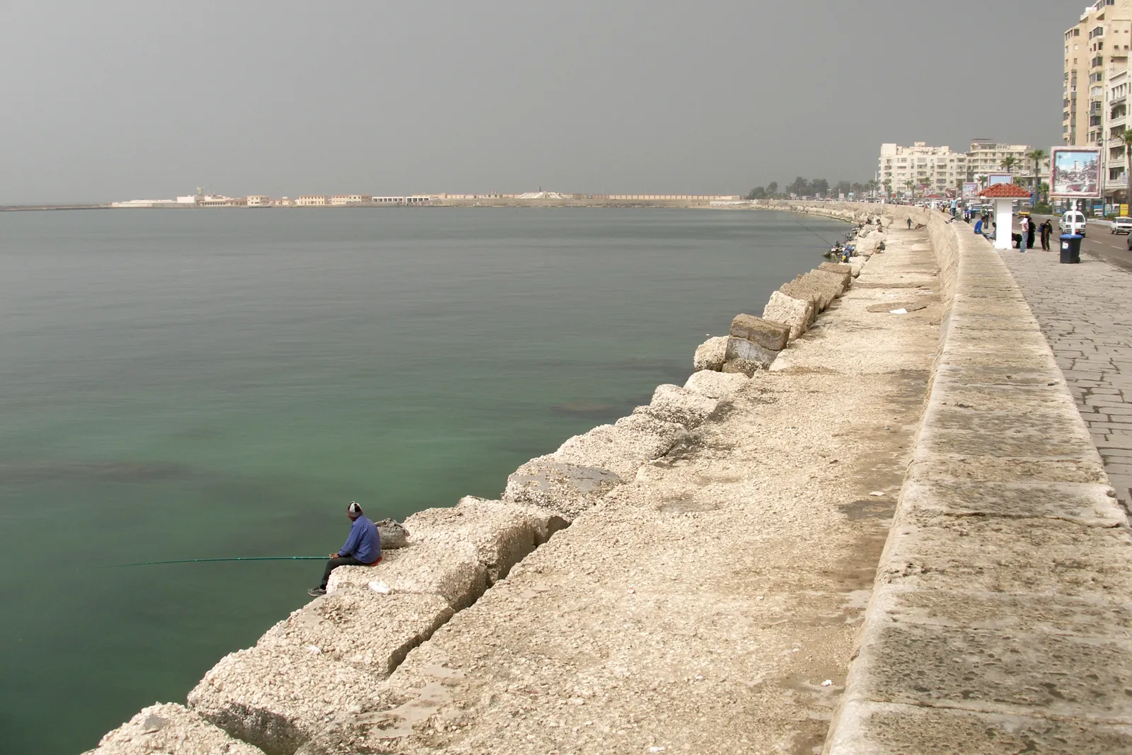 A man fishing with a rod from a stone seawall along a coastal promenade.