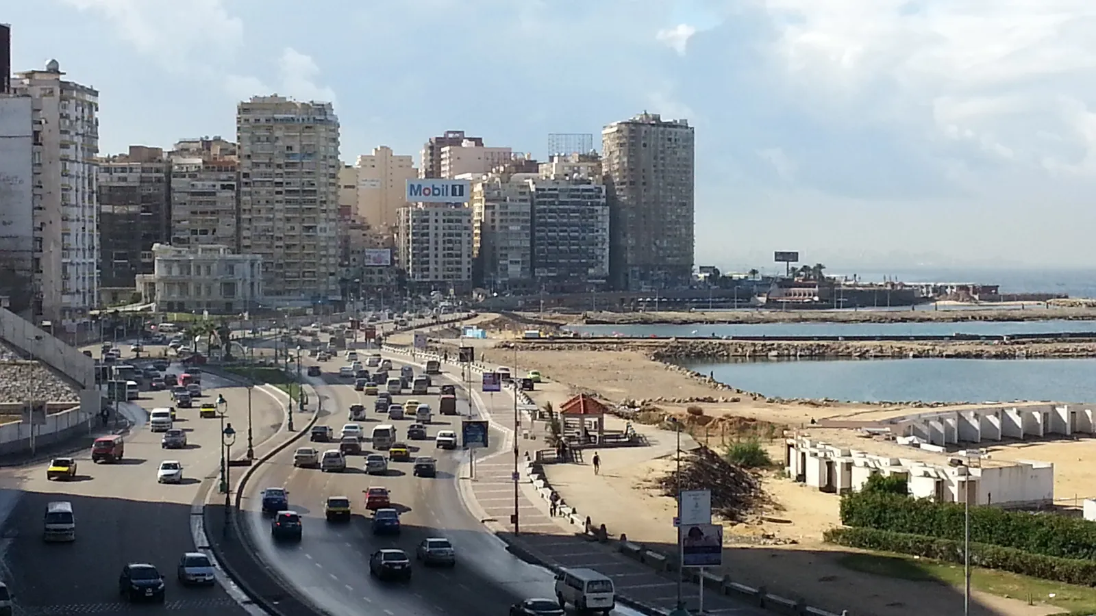 An aerial view of Alexandria's curved corniche road and beach lit by an orange sunset over the sea.