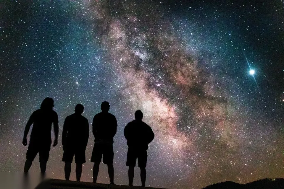 Four people stand in silhouette on a ridge watching the Milky Way arc across a star-filled desert sky.