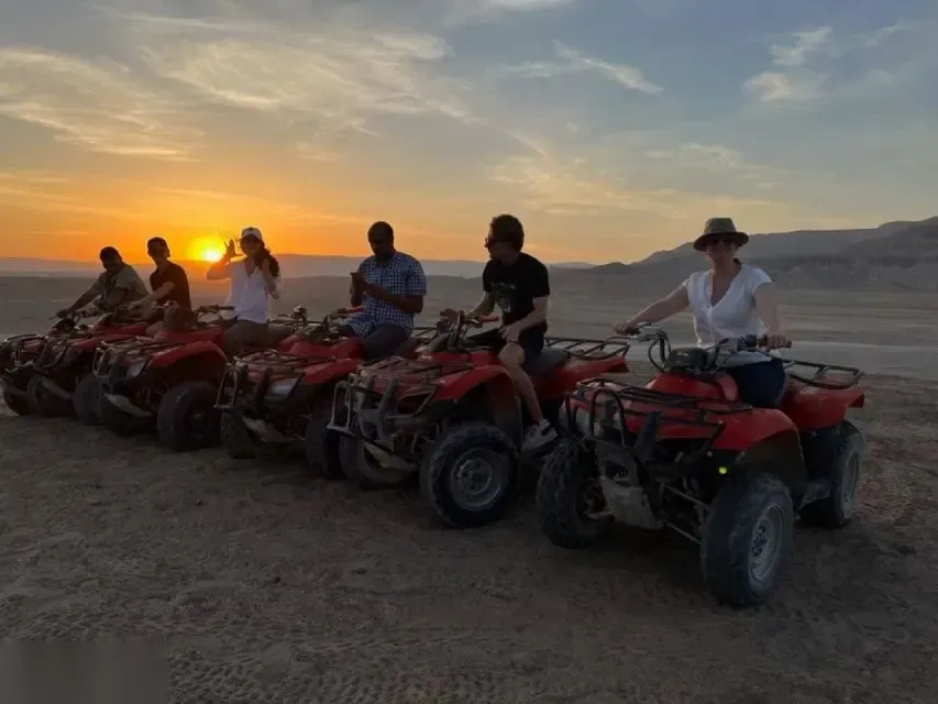 Six riders on red ATV quad bikes lined up in the desert as the sun sets behind distant hills.