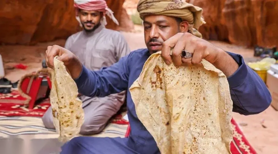 Two Bedouin men in a rocky desert camp hold up freshly baked flatbread for visitors to see.