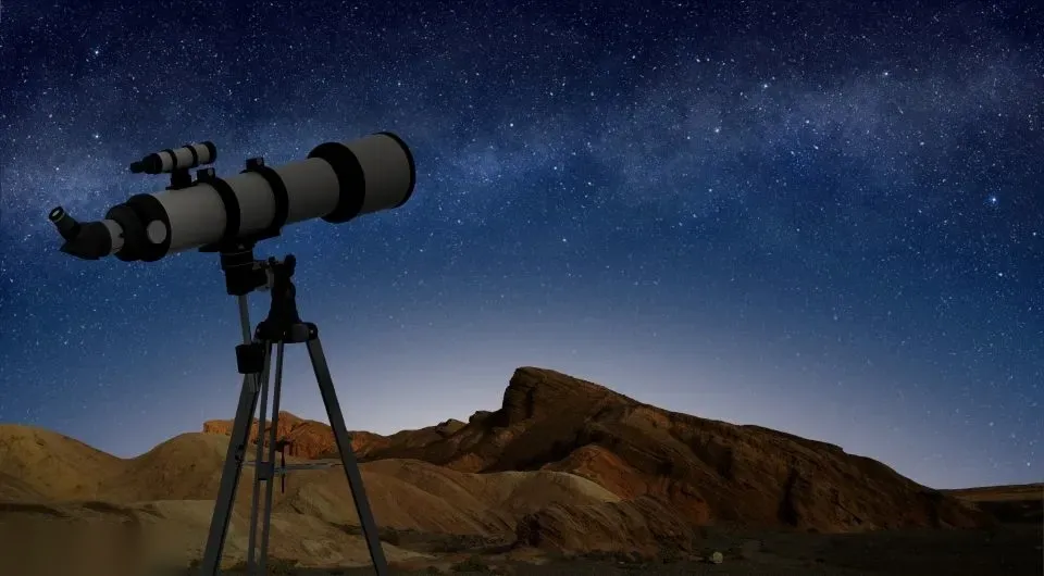 A telescope on a tripod is silhouetted against a starry night sky above rocky desert hills.