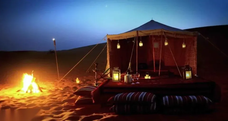 A lantern-lit Bedouin tent with cushions, a water pipe, and drinks set out beside a campfire at dusk.