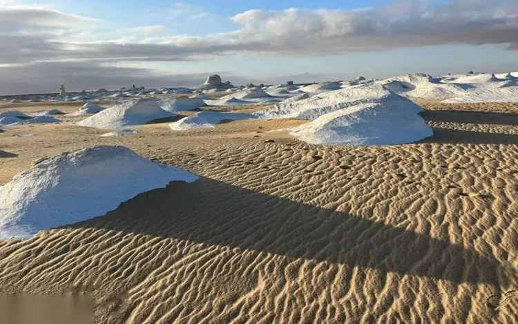 Snow-like white mineral mounds sit on rippled sand dunes under a partly cloudy sky at golden hour.