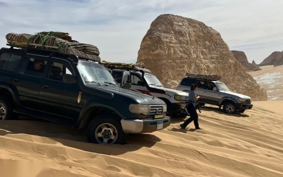 Three 4x4 Land Cruisers loaded with mattresses are parked beside a large boulder as a guide walks past.