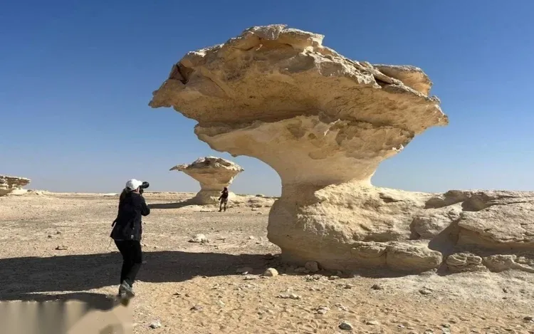 A photographer with a camera photographs a large mushroom-shaped white rock formation while another visitor looks on.