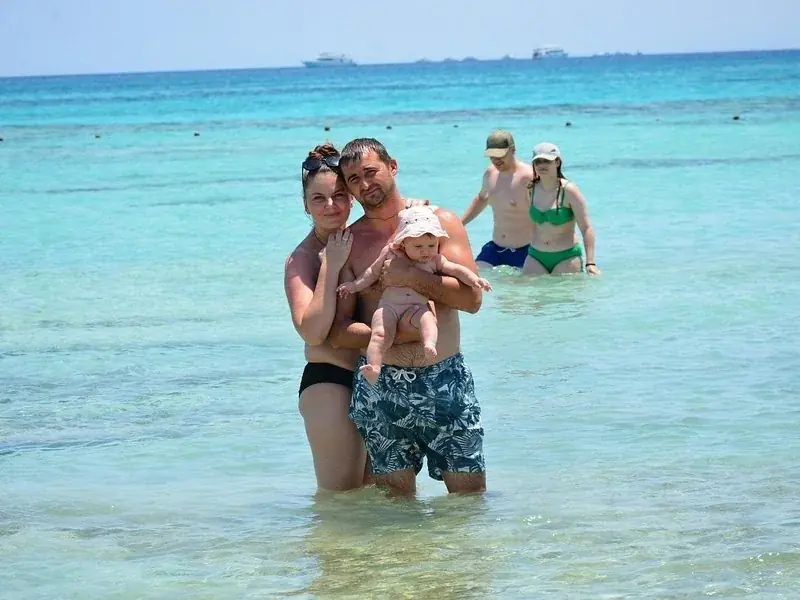 A family wades in shallow turquoise water, with the father holding a baby, while another couple stands in the background.