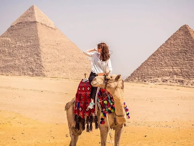 A tourist sits on a camel photographed from the side near the Great Pyramid in hazy desert light.