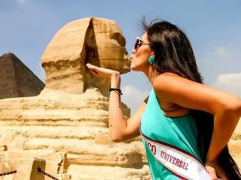A woman wearing a 'Miss Universe' sash blows a kiss toward the Great Sphinx, with a pyramid visible behind her.