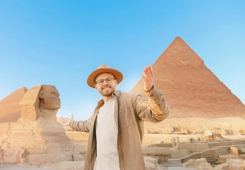 A man in a wide-brimmed hat smiles and gestures toward the Great Sphinx and Great Pyramid behind him.