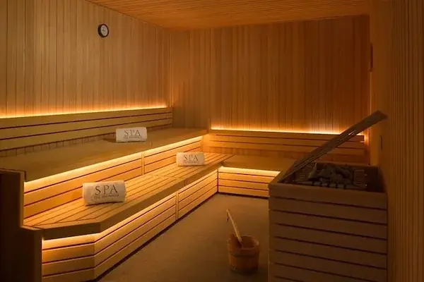 A wood-paneled sauna room with illuminated tiered benches holding folded towels labeled SPA, and a stove with a wooden bucket in the corner.