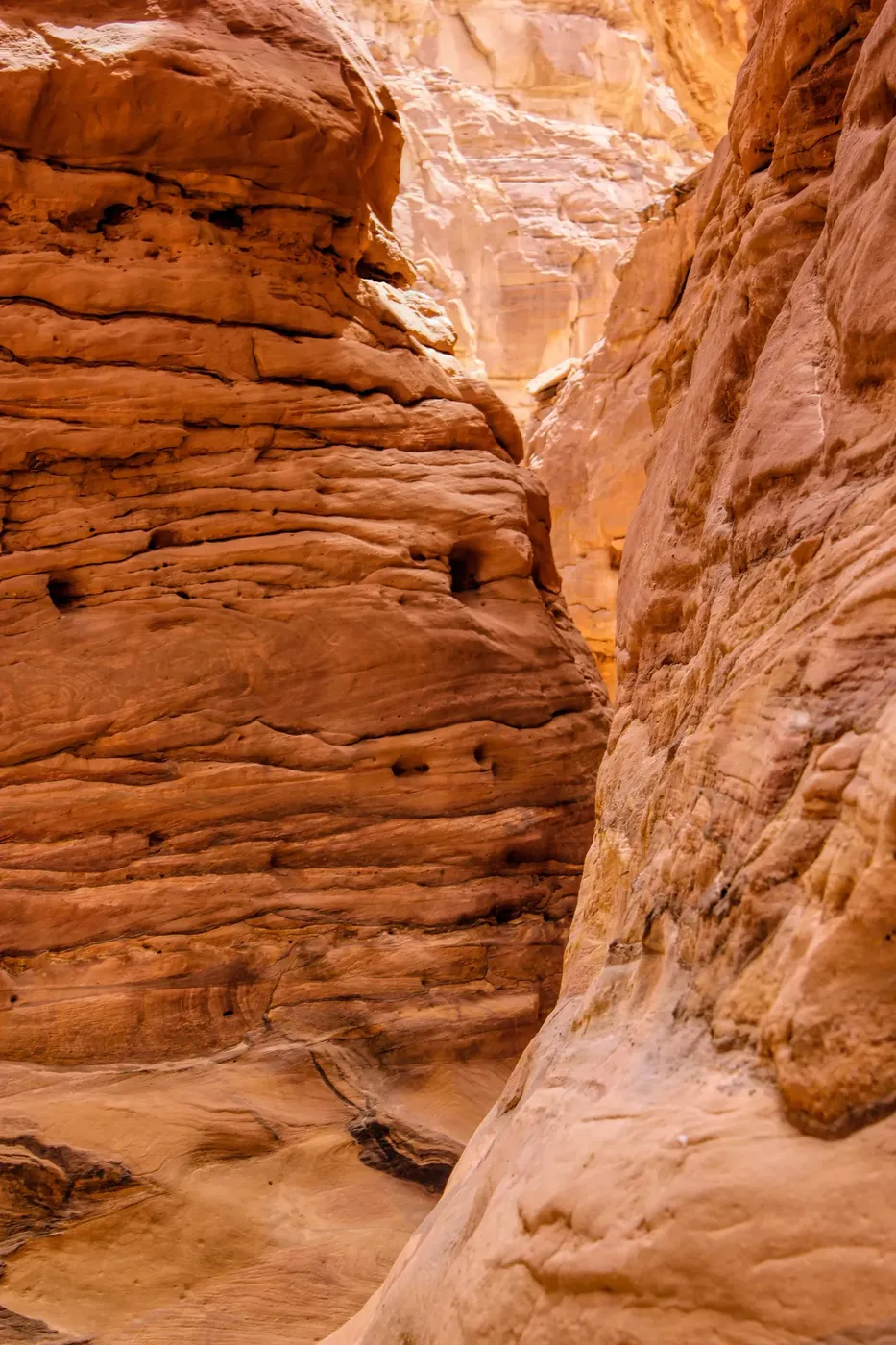 A long line of tourists in sun hats walks through a wide, sandy corridor between tall orange sandstone canyon walls.