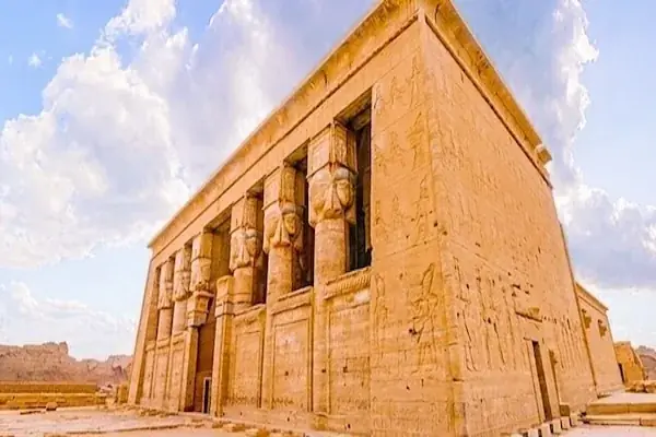 Dendera Temple rises behind a lush green field of crops with a palm tree at dusk.