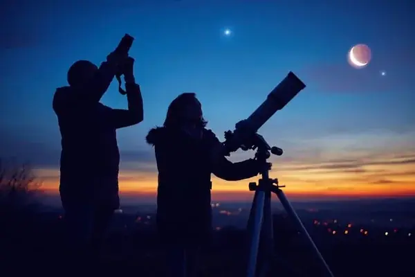 Silhouettes of two people using a telescope and camera to stargaze at dusk with a crescent moon visible.