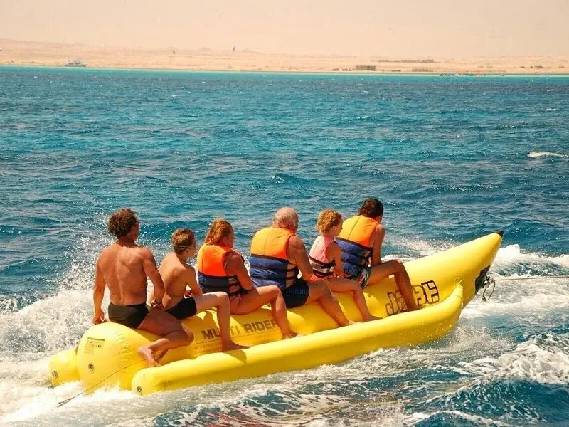 Family of six in orange and blue life vests riding a yellow banana boat towed across the sea, desert coastline behind them.