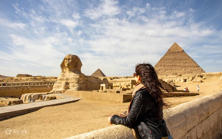 A line of ten tourists ride camels with arms raised in celebration, two pyramids visible in the background.