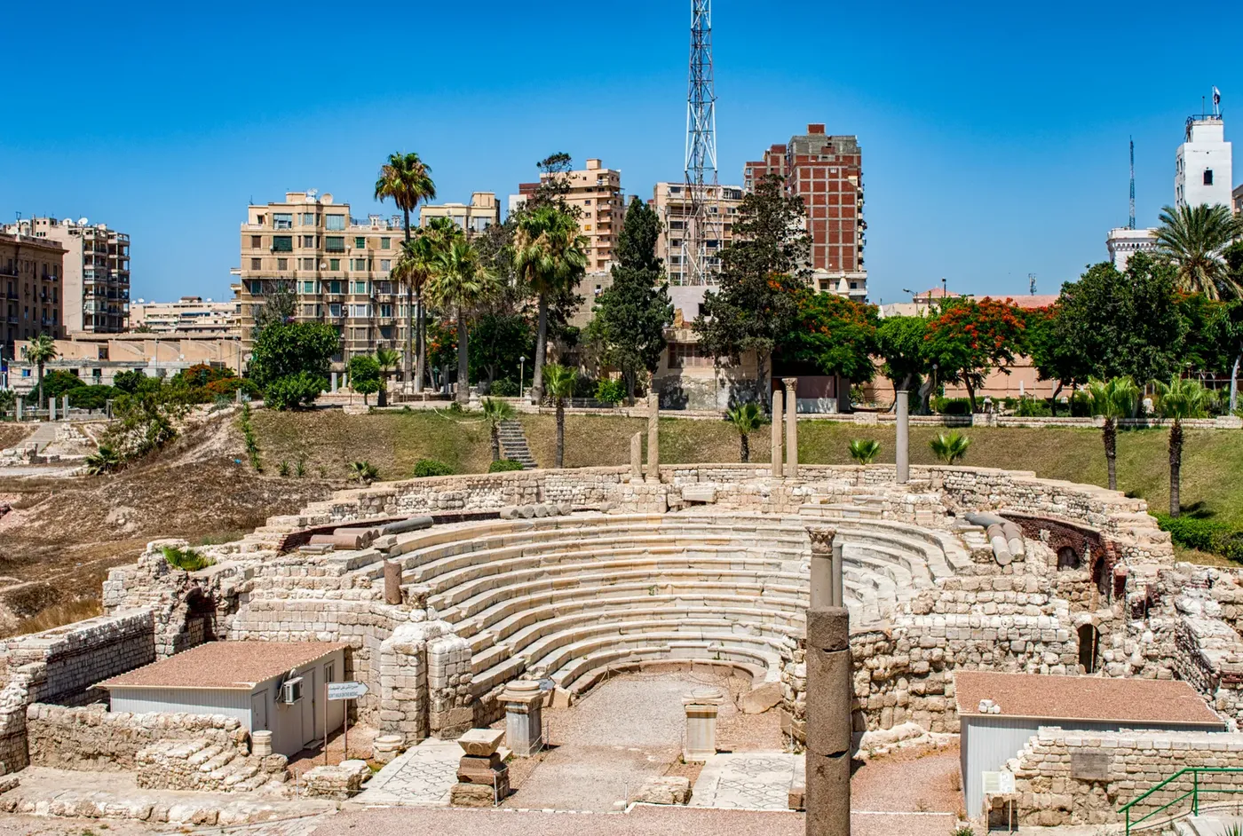 Pompey's Pillar, a tall red granite Roman column, stands above ancient ruins with two stone sphinxes at its base in Alexandria.