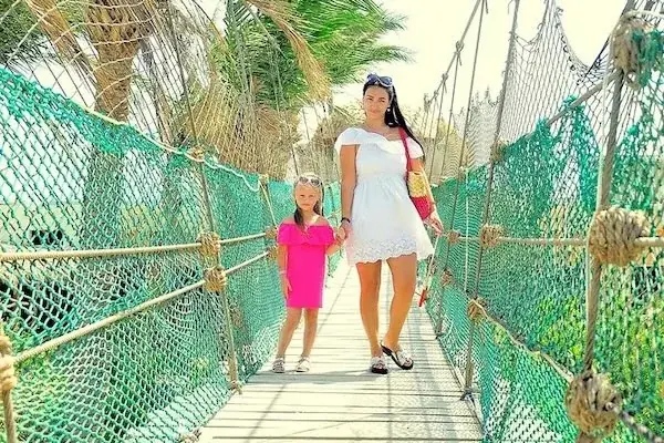 A mother and daughter walk hand in hand across a green rope suspension bridge lined with palm trees.