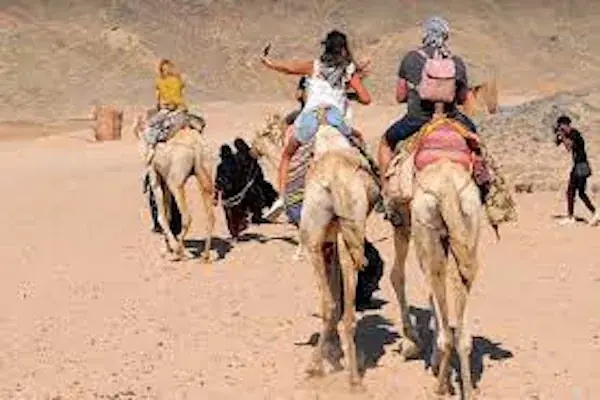 Tourists riding camels through the desert, one taking a selfie, with a guide on foot leading them.