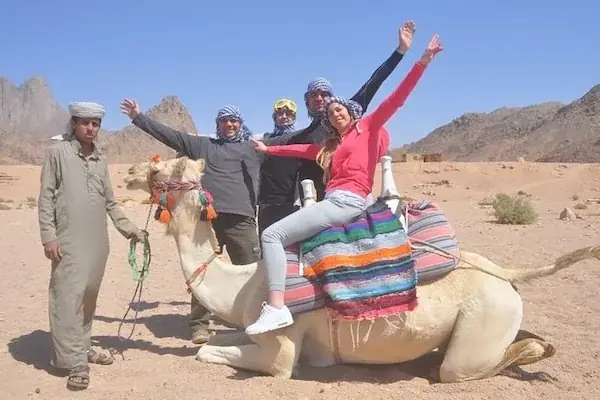 A group of tourists posing with raised arms next to a seated camel and a Bedouin guide, with mountains behind.