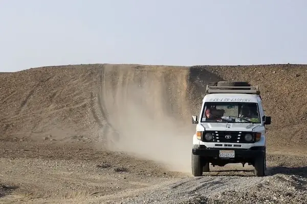 A white 4x4 jeep with two passengers driving along a dusty desert track, kicking up dust behind it.