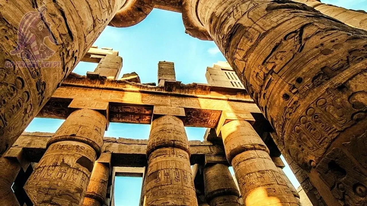 Tourists with an umbrella walk toward the towering sandstone pylon and obelisk of Karnak Temple in Luxor.