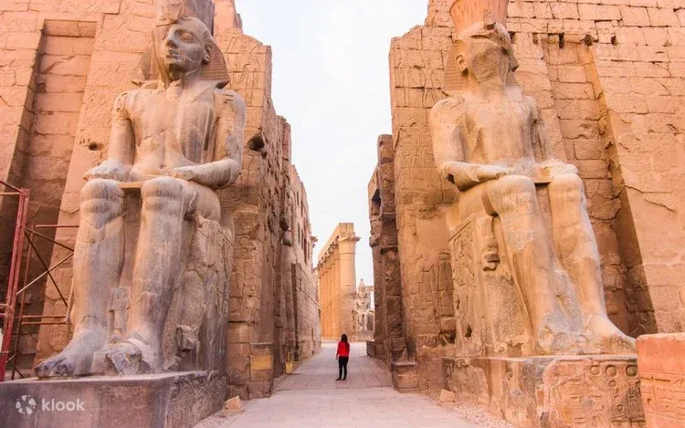 A large crowd of tourists in sun hats walks between massive stone pylons toward a distant obelisk at Karnak Temple.