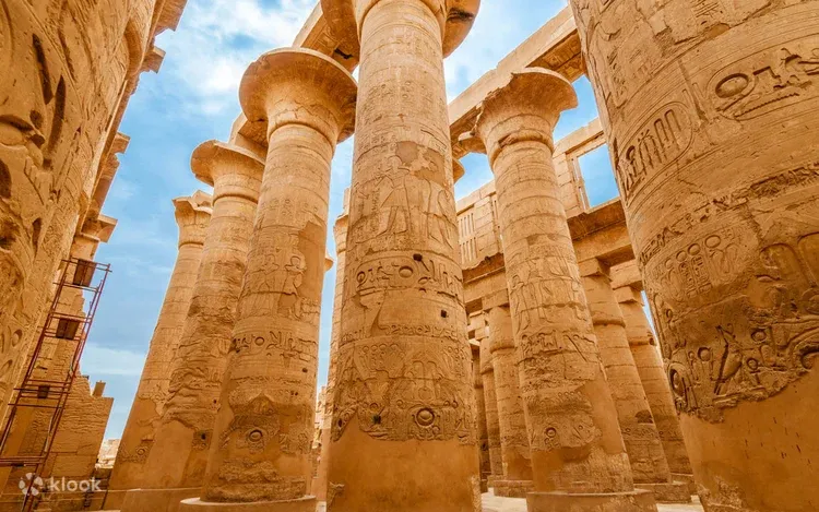 A woman in a blue cap and backpack poses in front of ancient statues and columns at Karnak Temple under a blue sky.