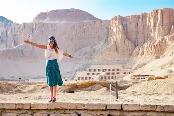 A woman in a teal skirt and headscarf poses with arms outstretched on a wall overlooking Hatshepsut Temple and the desert cliffs.