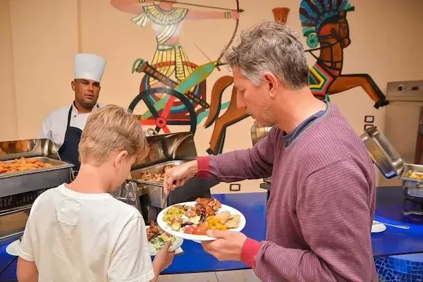 A chef serves a father and son from a buffet, with a colorful pharaonic-style mural painted on the wall behind them.
