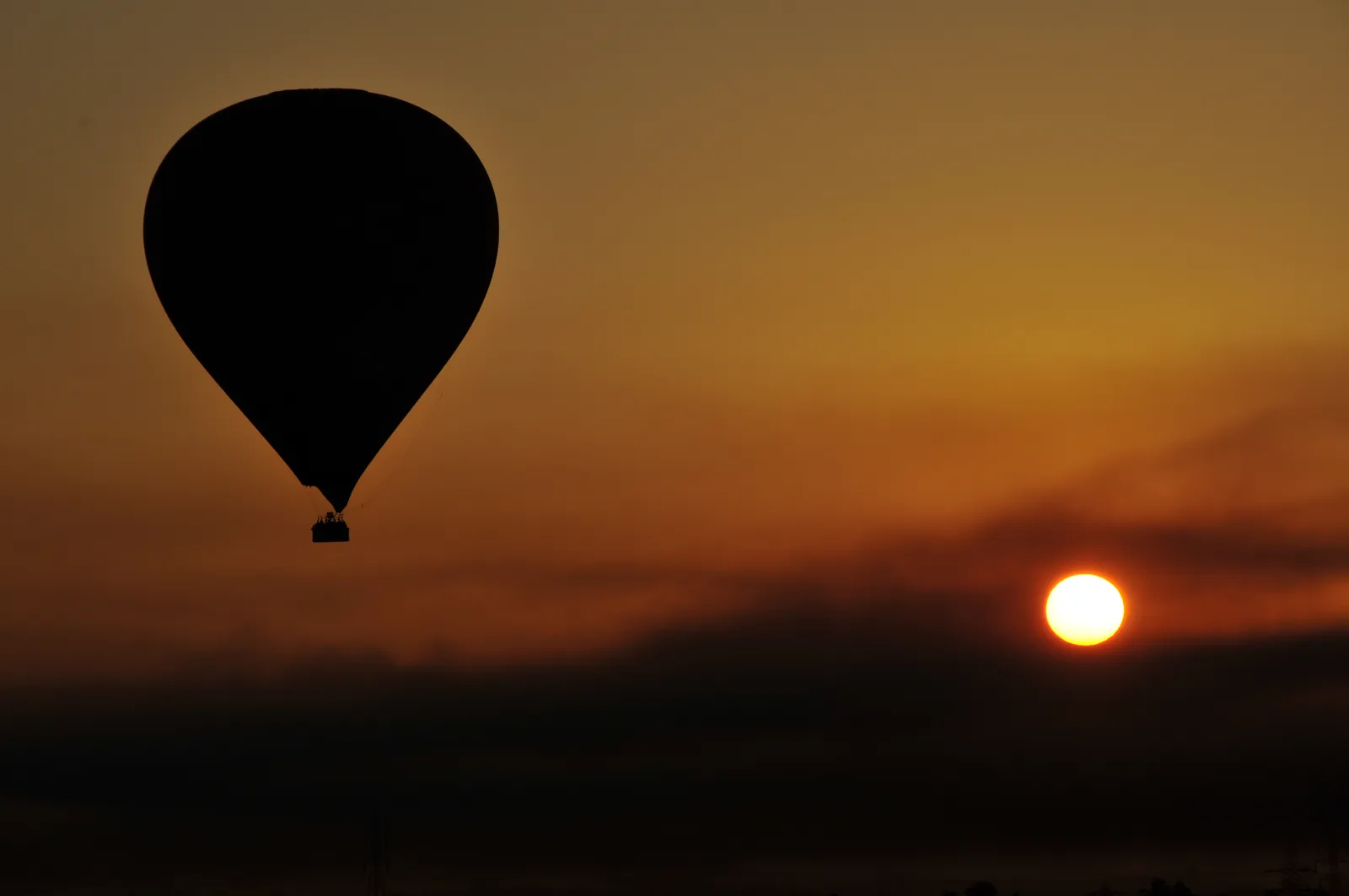 A large rainbow-striped hot air balloon flies over the Nile River and Luxor's temple ruins, with several other balloons in the sky.