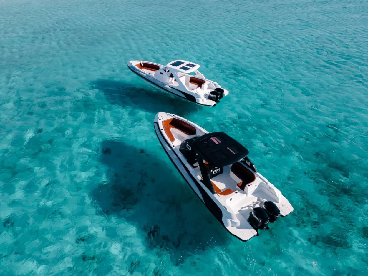 Tourists on an inflatable speedboat lean over to watch a dolphin swimming beside them in clear turquoise water, another boat and yacht in the background.