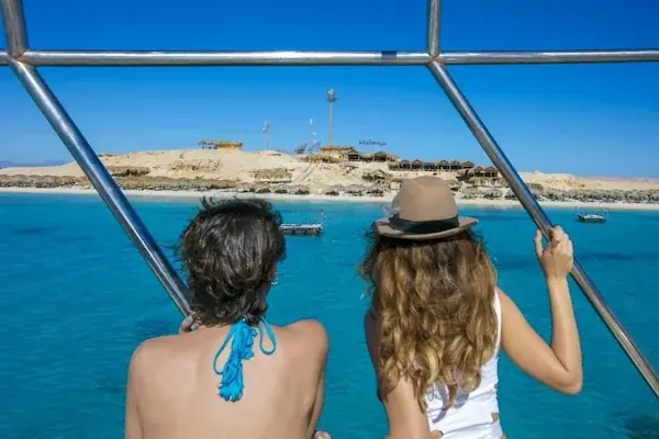 Straw beach umbrellas on golden sand with several tour boats anchored in the turquoise bay and hazy mountains beyond.