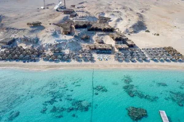The Mahmya sign and thatched huts on a sandy cliff above a crowded beach, seen from a passing boat.