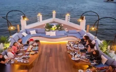 Aerial view of guests seated at curved sofa tables on the boat's bow, dining as evening lights come on over the river.