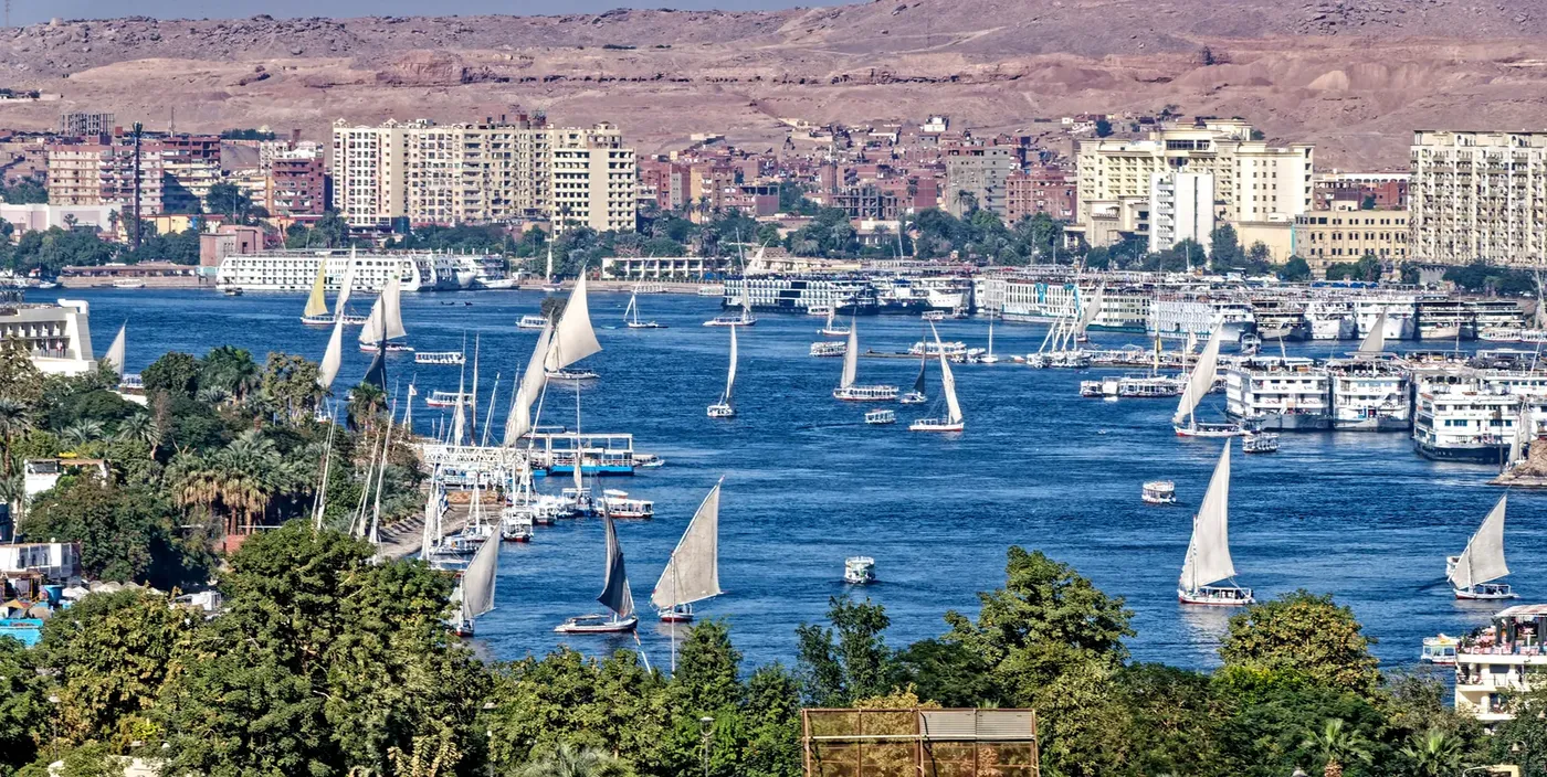 A cluster of tall Nubian houses in blue, yellow, and pink stacked up a rocky slope above the Nile, with boats moored below.