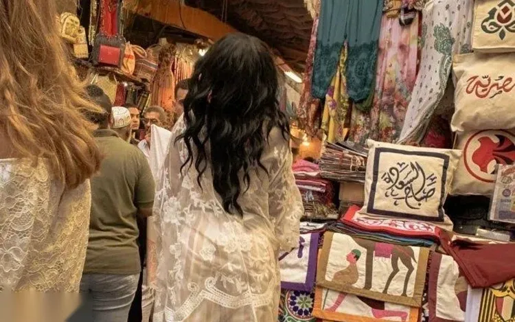 A woman in a patterned jumpsuit walks through a Khan el-Khalili alley lined with hanging lanterns, brass ornaments, and pottery stalls.