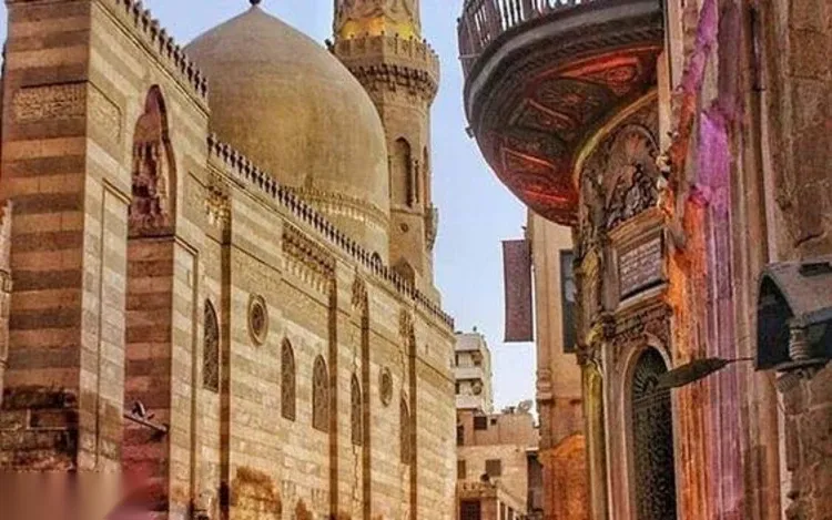 A narrow lantern-lined alley in Khan el-Khalili leads through a stone archway toward more market stalls, with a cat on the cobblestones.