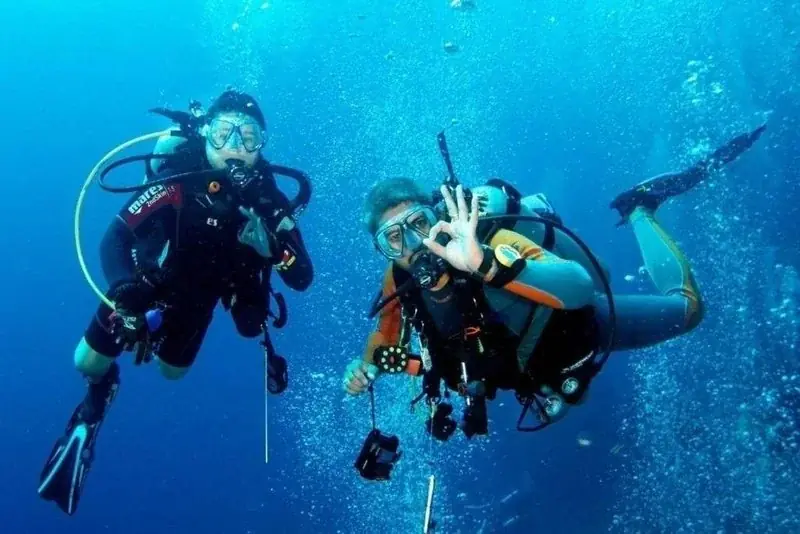 Group of four students and an instructor sitting poolside with dive gear laid out in front of them, resort loungers and shade structures behind.