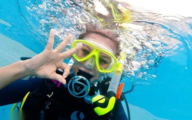 A diver in a yellow mask gives an OK hand sign in a selfie taken just under the surface.