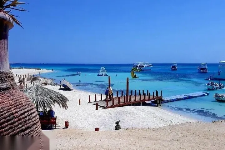 A large blue PARADISE sign stands on the beach above straw cabanas and a crowded turquoise swimming area.