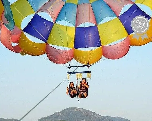 A multicolored parasail canopy carries two riders high above the sea while a crew member on the boat holds the tow line.