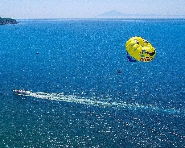 Looking straight up at a bright blue parasail canopy with two riders below, arms outstretched.