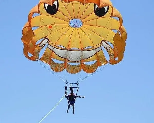Two riders in life vests grin and wave from a parasailing harness against a clear blue sky.