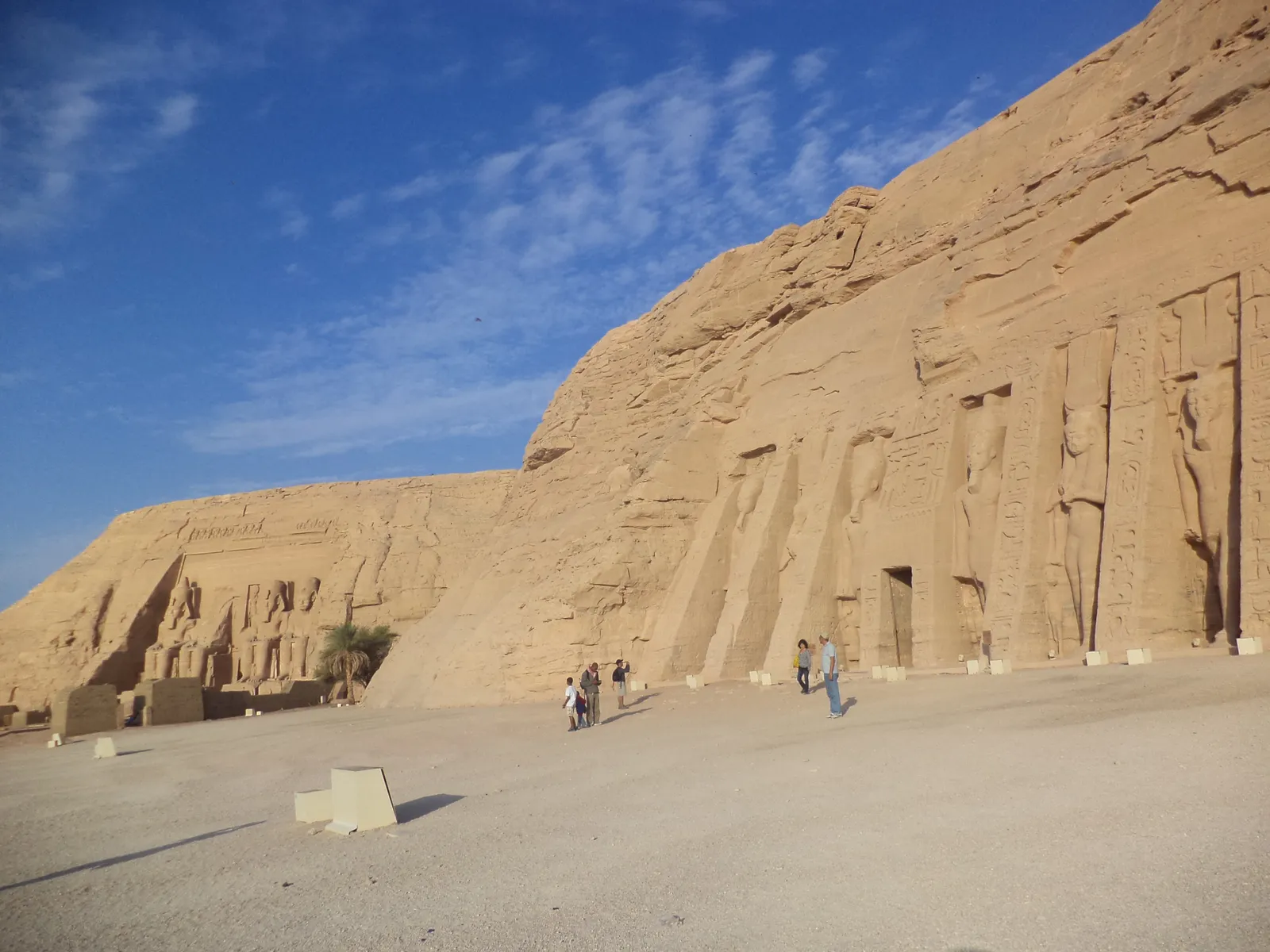 Rows of tour coaches parked in a desert lot near Abu Simbel, with a low sandy hill and palm trees in the background.