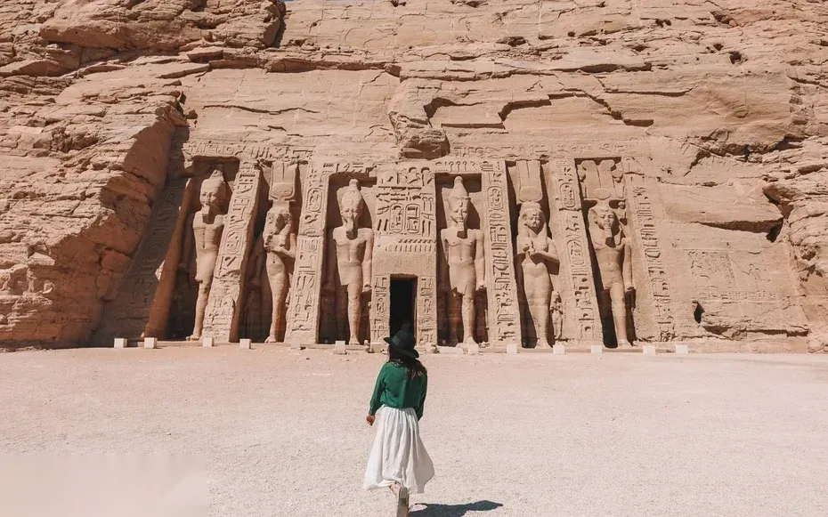 A woman in a green top and white skirt walks toward the carved facade of the Temple of Hathor at Abu Simbel.