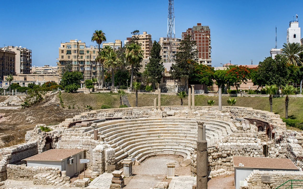 Couple standing among the weathered limestone tiers of the Roman Amphitheatre (Kom el-Dikka) with ancient columns and palm trees behind them
